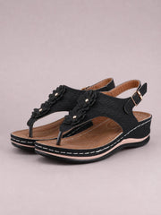 Black toe post flower detail wedge sandals feature floral appliqués, perforated straps, adjustable ankle buckles, tan footbeds, and mid-height wedge soles.