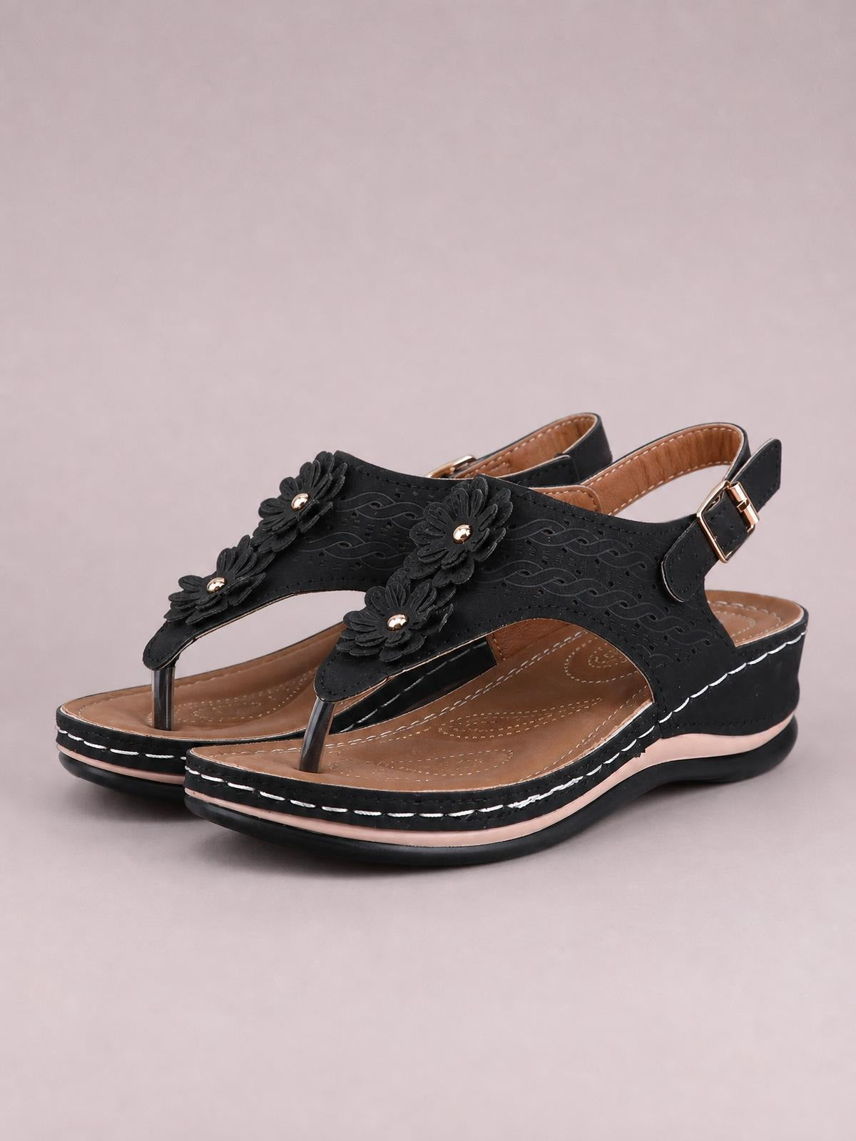 Black toe post flower detail wedge sandals feature floral appliqués, perforated straps, adjustable ankle buckles, tan footbeds, and mid-height wedge soles.