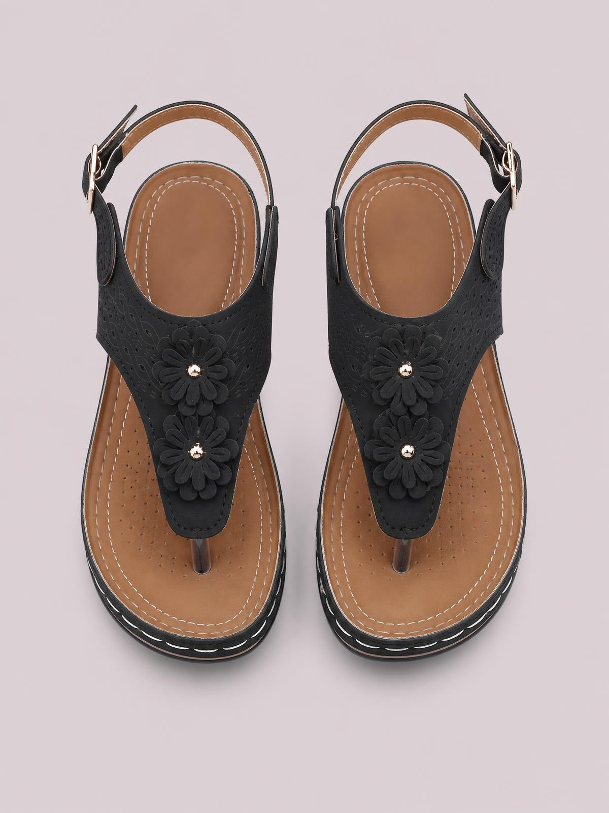 Black toe post wedge sandals feature layered flower details, perforated uppers, ankle straps with buckles, tan insoles, and contrasting stitched soles.