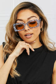 Model poses with one hand beneath her chin, wearing bold rectangle polycarbonate sunglasses with patterned tortoiseshell frames and amber lenses.