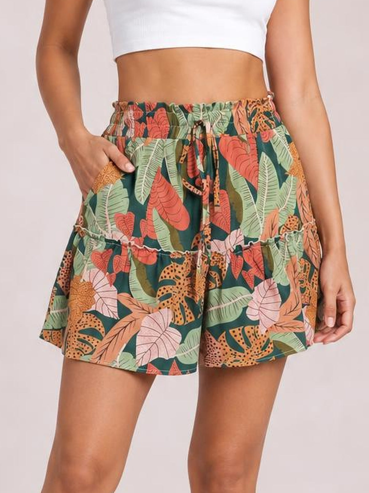 Model wears tropical print high waist shorts in dark teal with leafy florals and leopard motifs, a tied elastic waist, side pockets, and a frilled hem.