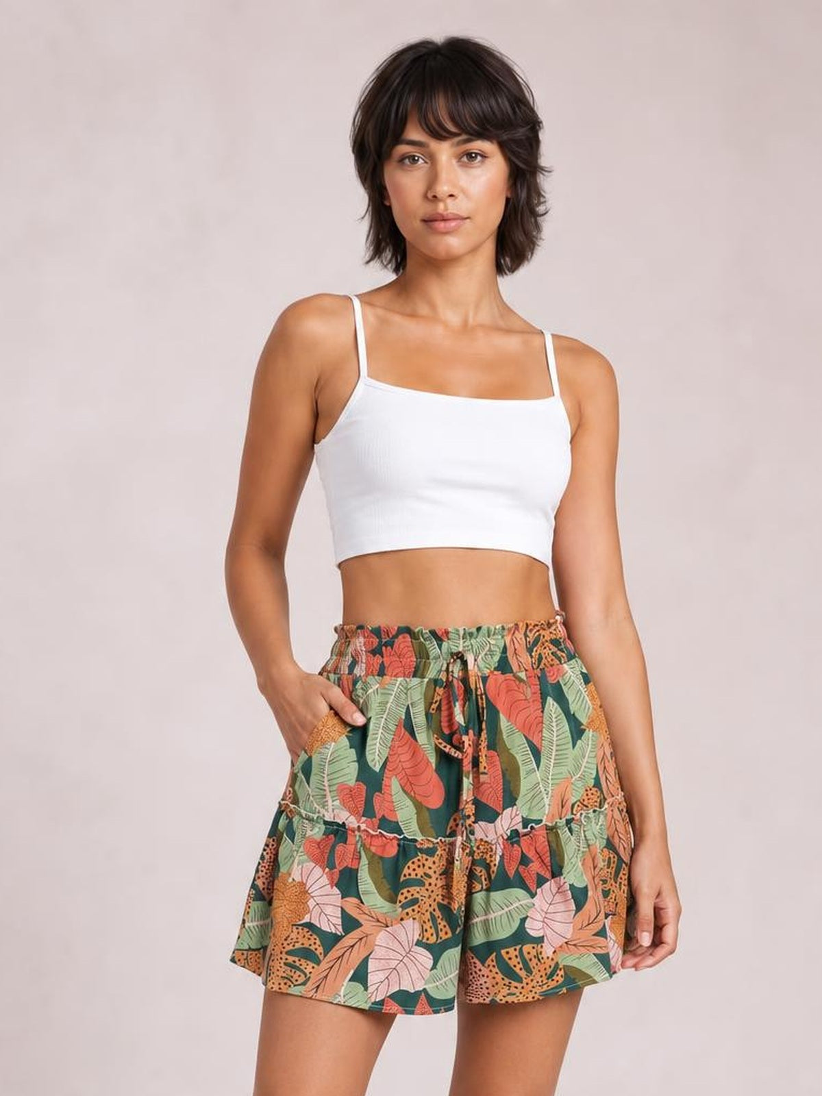A woman modeling a white crop top and high-waisted shorts with a colorful tropical leaf print in green, orange, and pink, set against a solid light beige background.