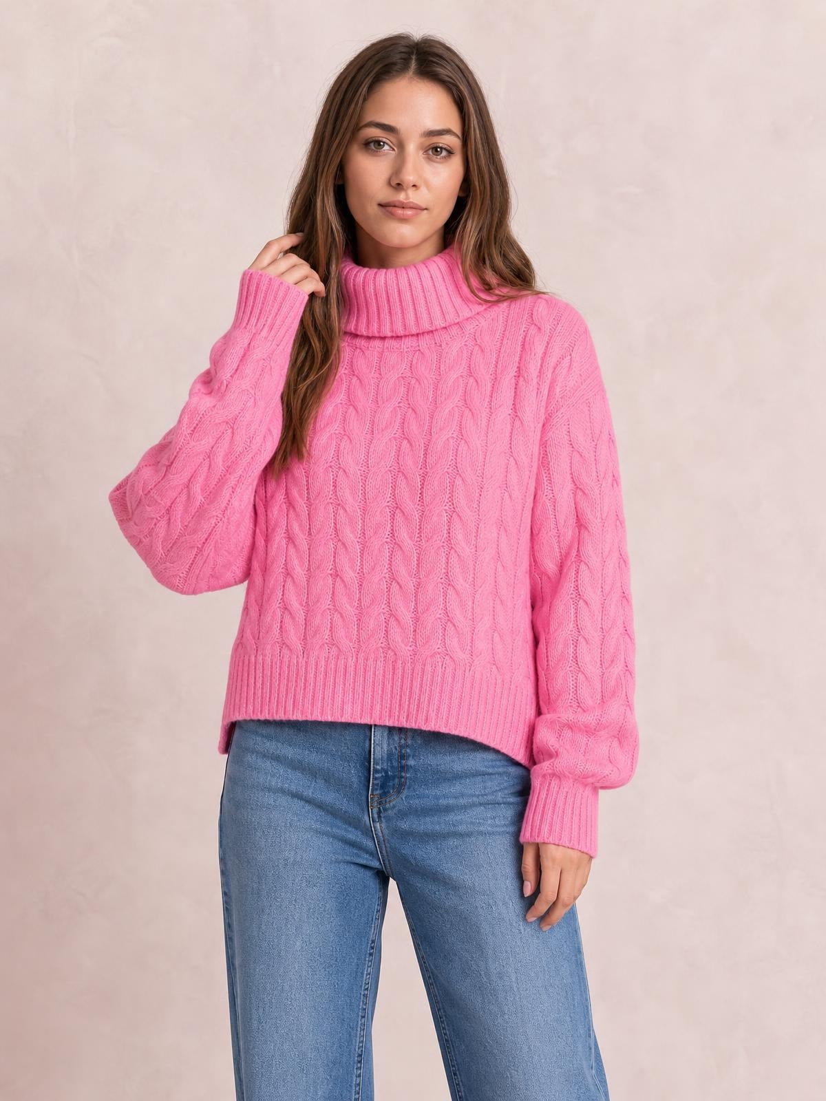 Model poses in a bright pink cable-knit turtleneck sweater with long sleeves, ribbed cuffs and hem, and a regular-length relaxed silhouette.