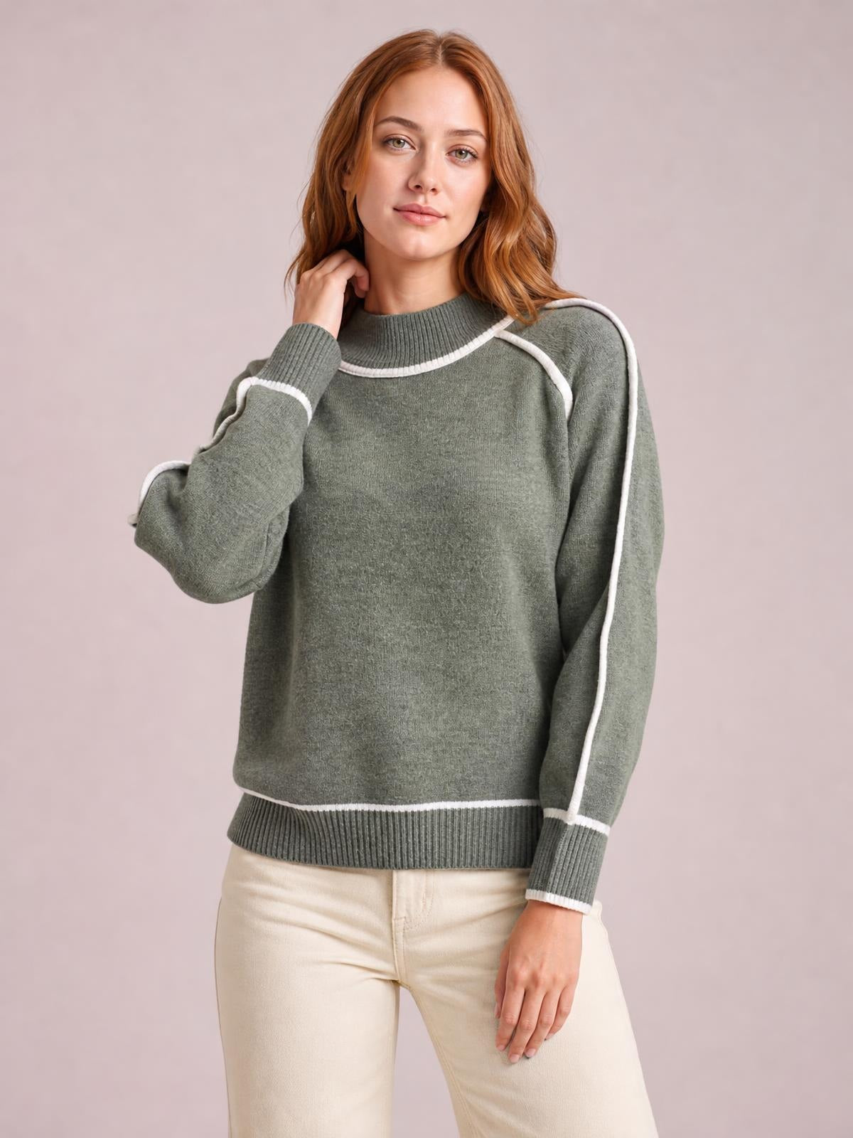Model wearing a relaxed olive-gray turtleneck sweater with white contrast trim, ribbed collar, cuffs and hem, posing with one hand near her neck.