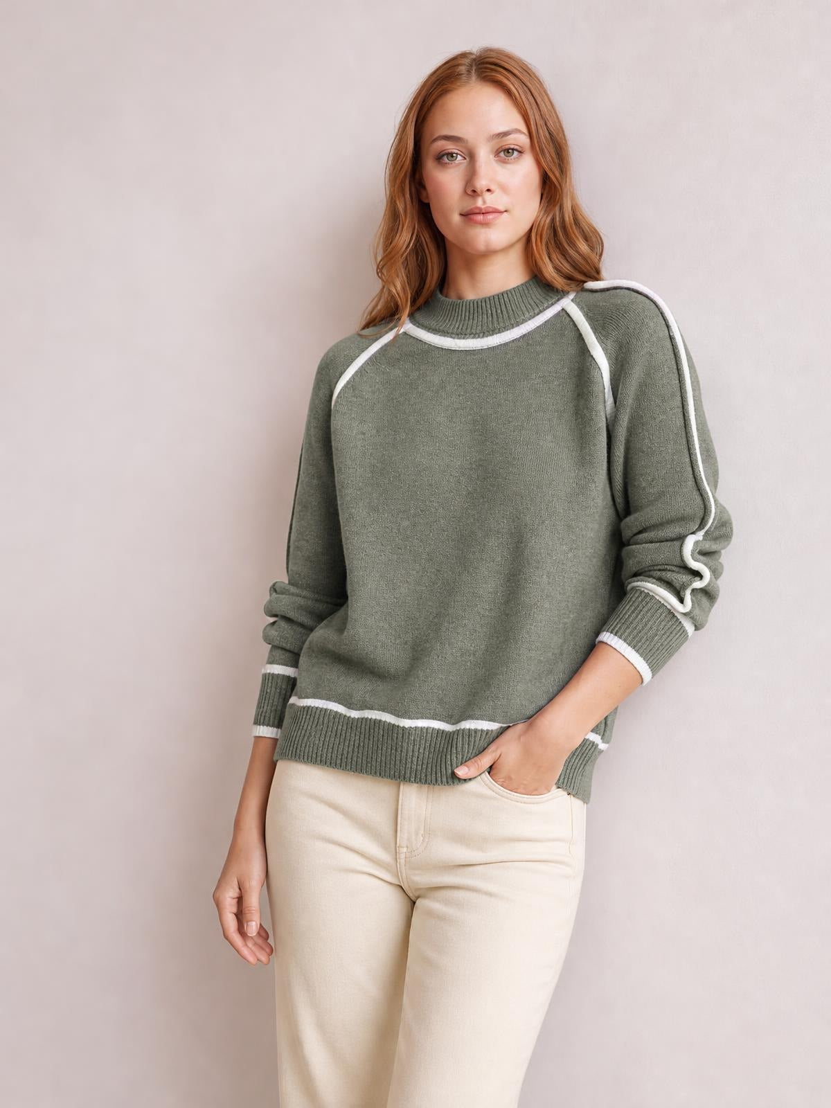 Model wears a relaxed olive-green turtleneck sweater with white contrast trim, ribbed collar, cuffs, hem, and sleeve details, posing with one hand in her pocket.