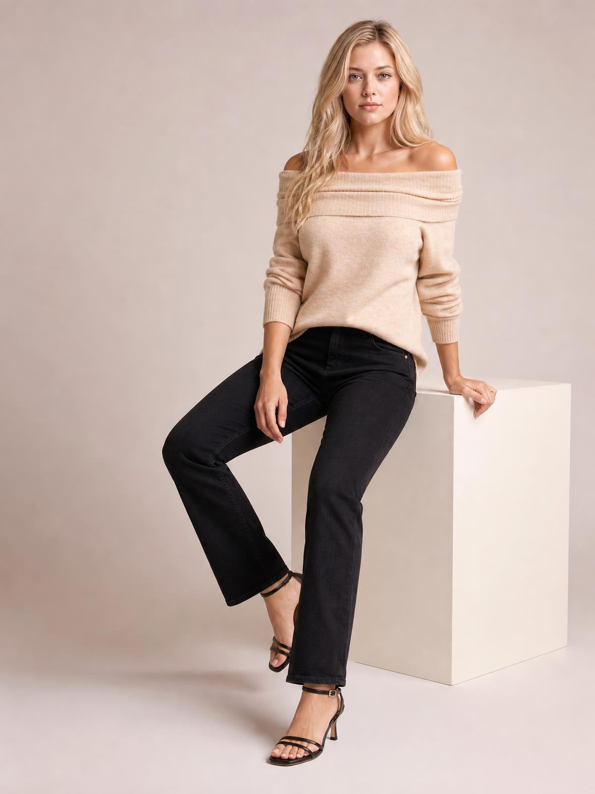 A model sits wearing a beige long-sleeve sweater with a wide folded neckline, relaxed silhouette, ribbed trim, and fitted cuffs.