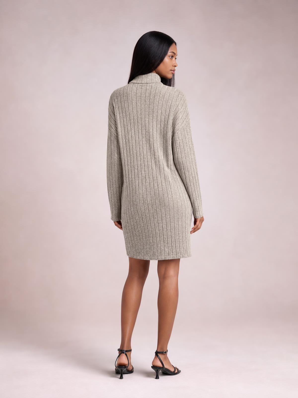 Model looks over her shoulder in a beige ribbed knit dress with a turtleneck, long sleeves, fitted silhouette, and above-the-knee hem.