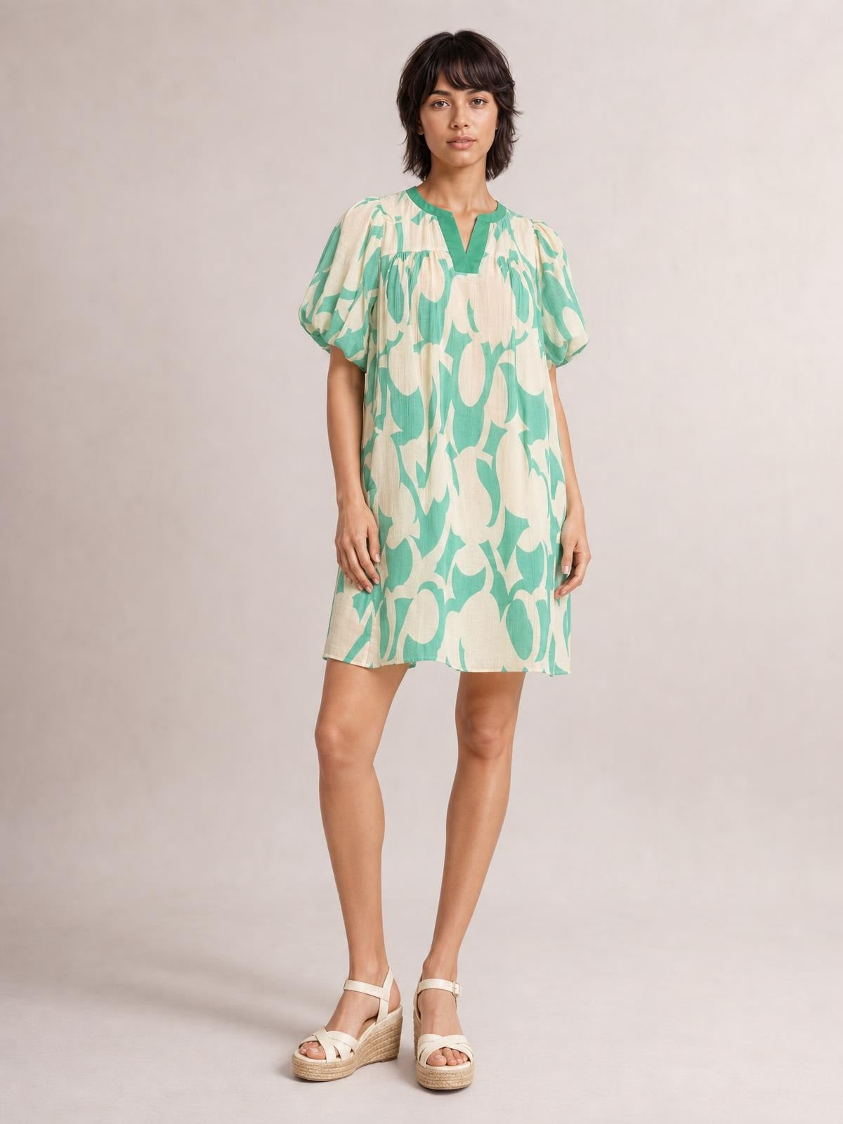 A model stands in a relaxed pose wearing a mint green and beige abstract-print mini shift dress with puff sleeves, a V-neckline, and a flowy silhouette.