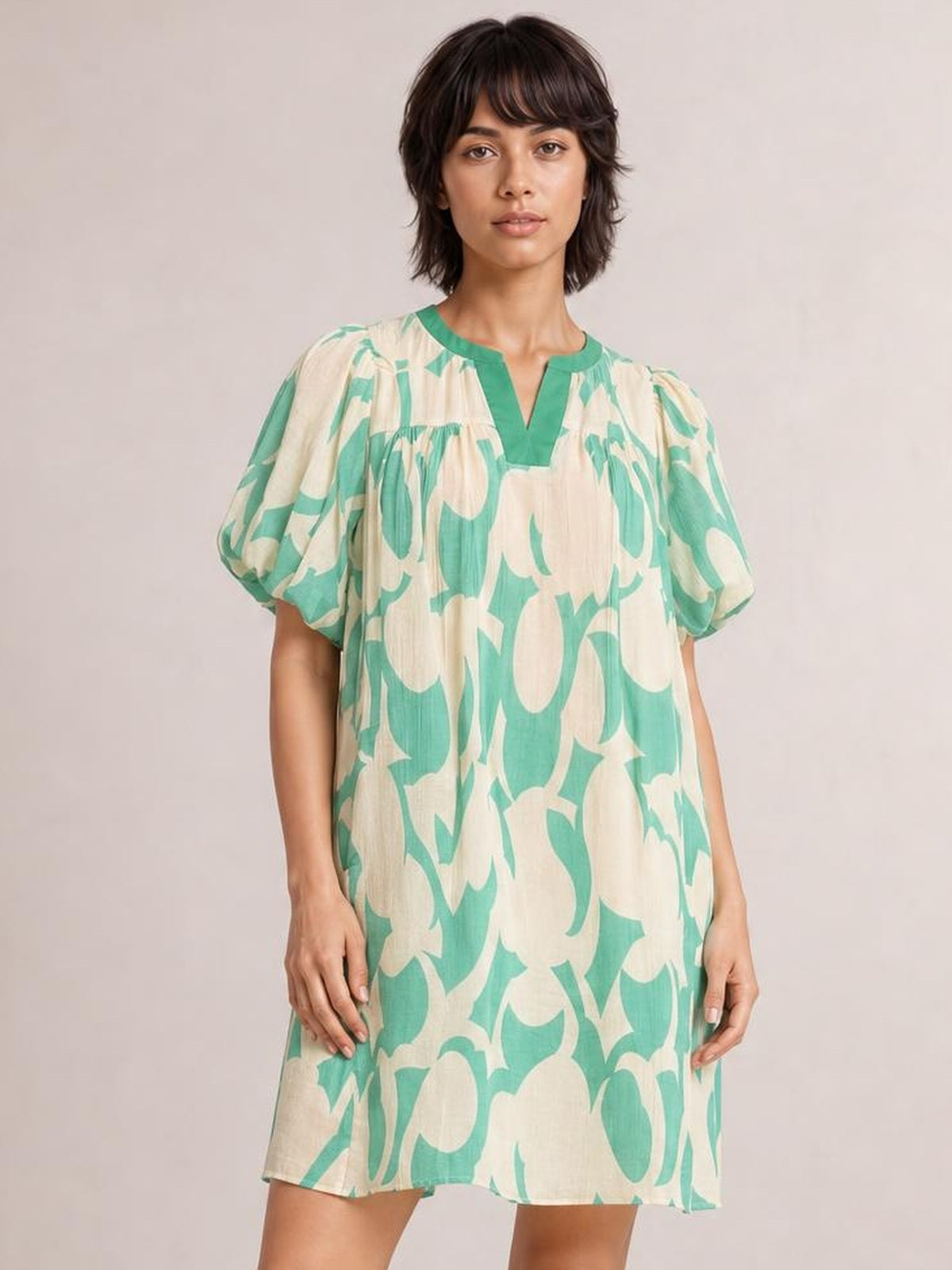 A model stands in a mint green and beige abstract print shift mini dress with puff sleeves, a split V-neckline, gathered shoulders, and a relaxed silhouette.