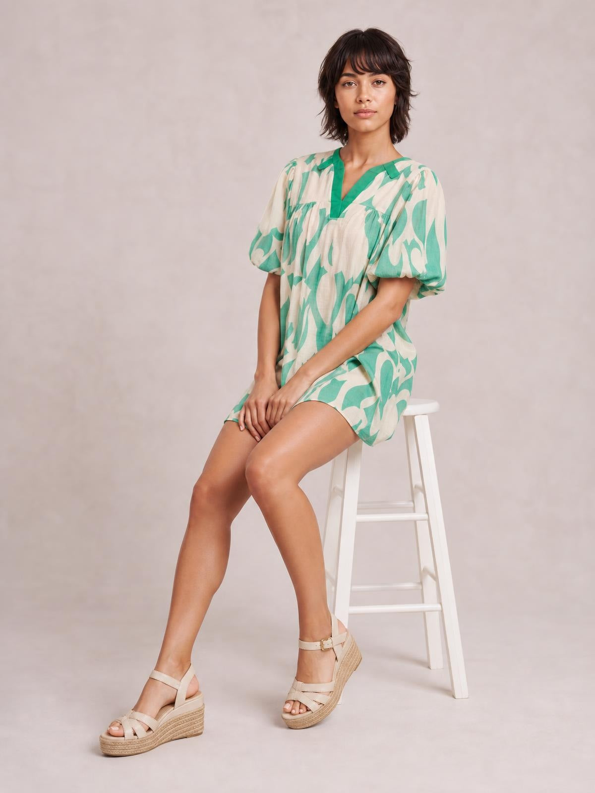 A seated model wears a mint green and beige abstract print mini shift dress with a V-neckline, puff short sleeves, and a relaxed flowy silhouette.