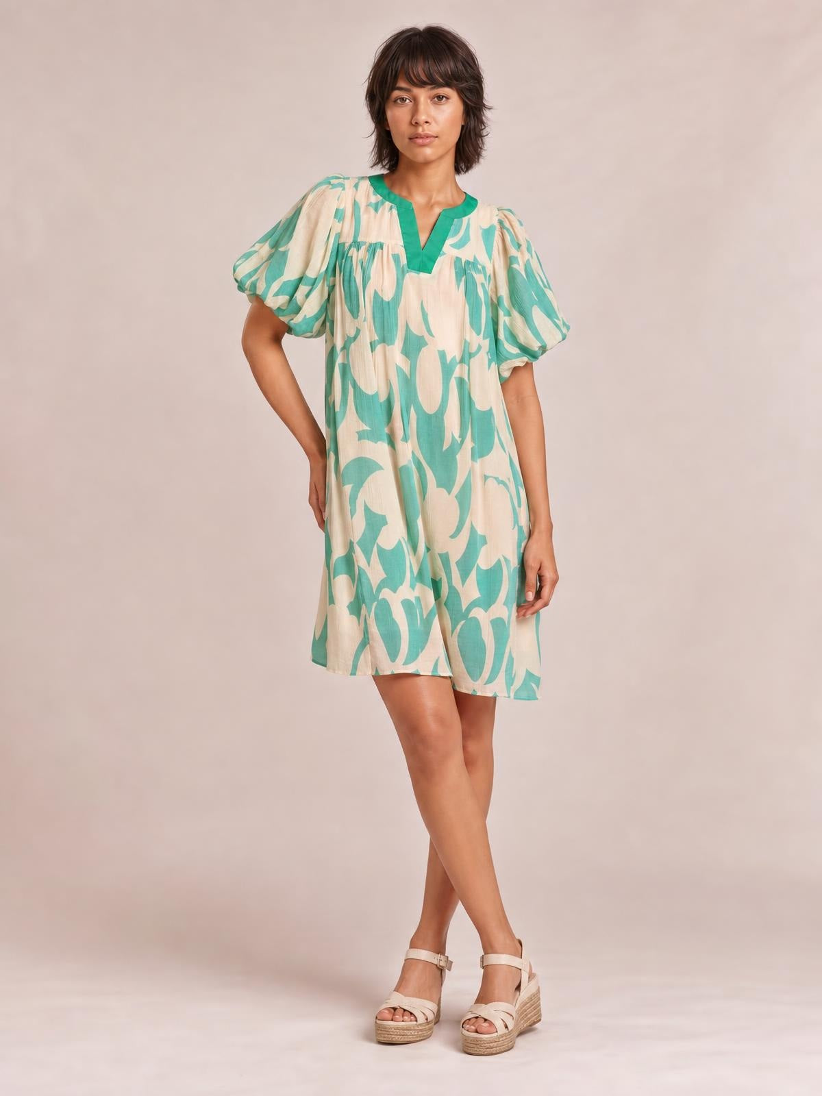 A model poses with crossed legs in a mint green and beige abstract-print mini shift dress featuring puff sleeves, a V-neckline, and a relaxed silhouette.