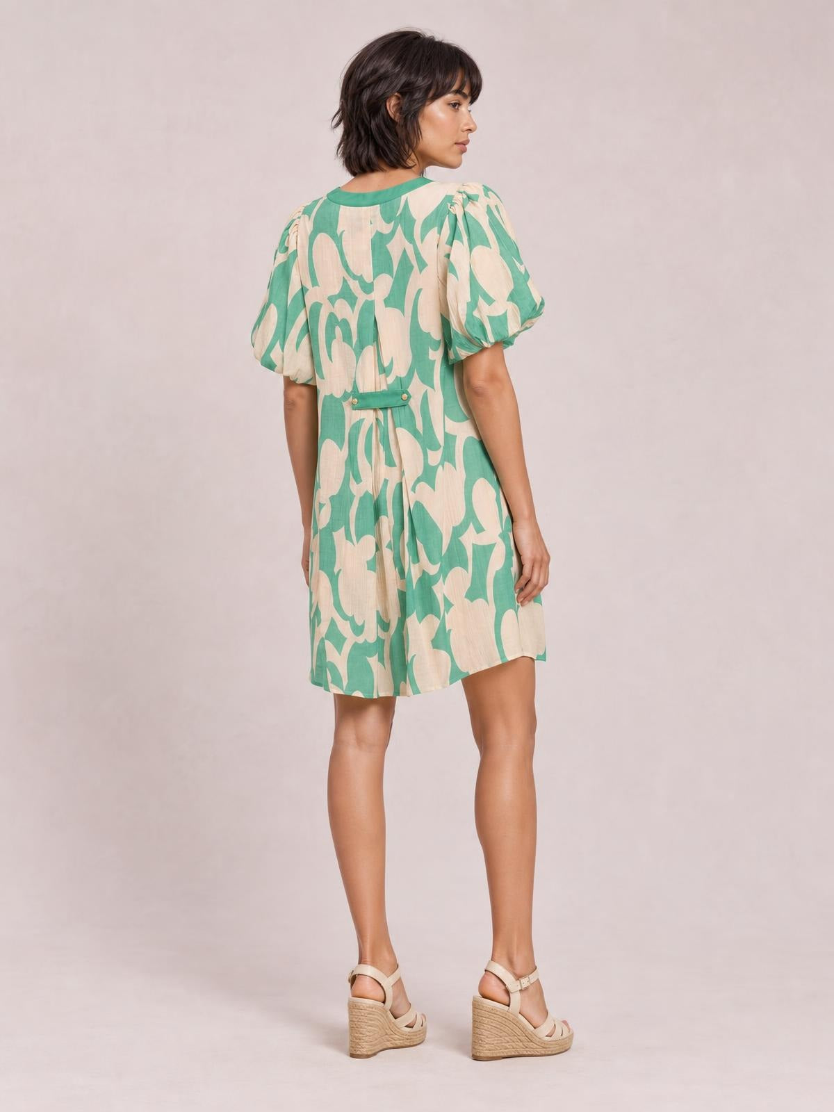 A model poses from the back in a mint green and beige abstract print mini shift dress with puff sleeves, a relaxed silhouette, and a buttoned waist tab.