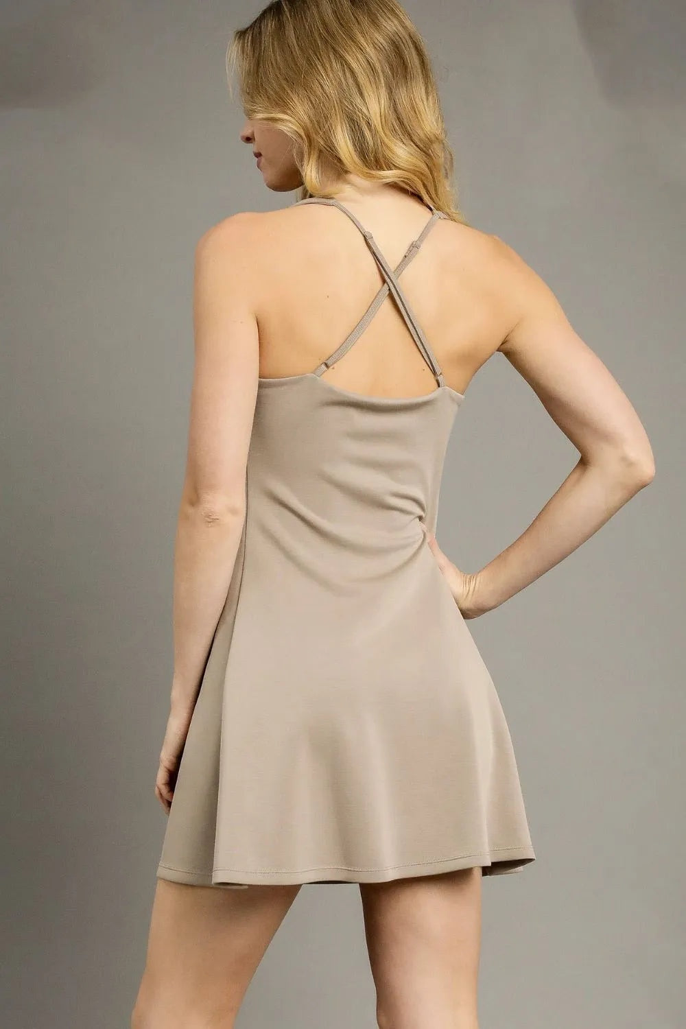 Model poses with one hand on her hip, showing the beige sleeveless active mini dress’s crisscross back straps and flowing A-line silhouette.