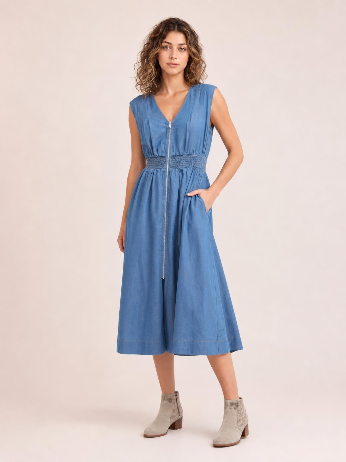 Model poses in a blue denim sleeveless midi dress with a V-neck zip front, smocked waist, side pockets, and a softly flared skirt.