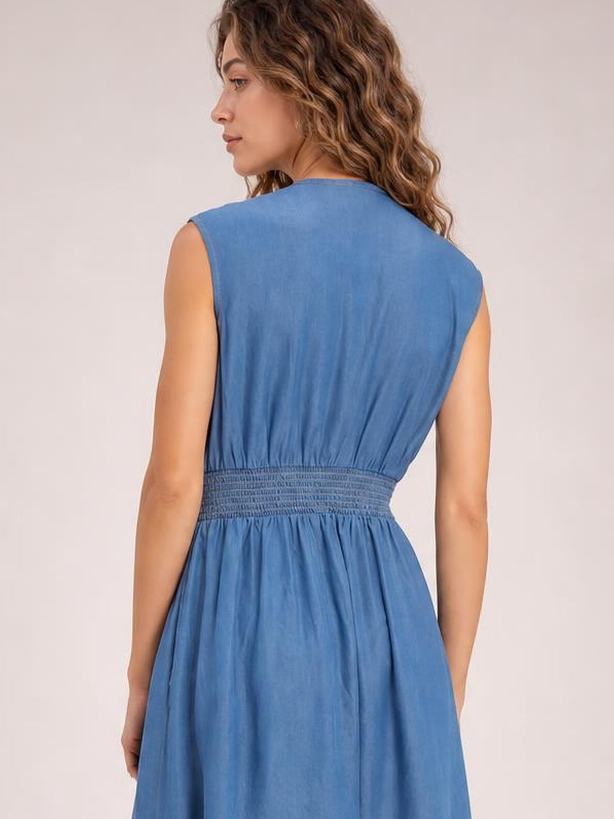 A model shown from behind wears a blue denim sleeveless midi dress with a fitted smocked waist and softly flared skirt silhouette.