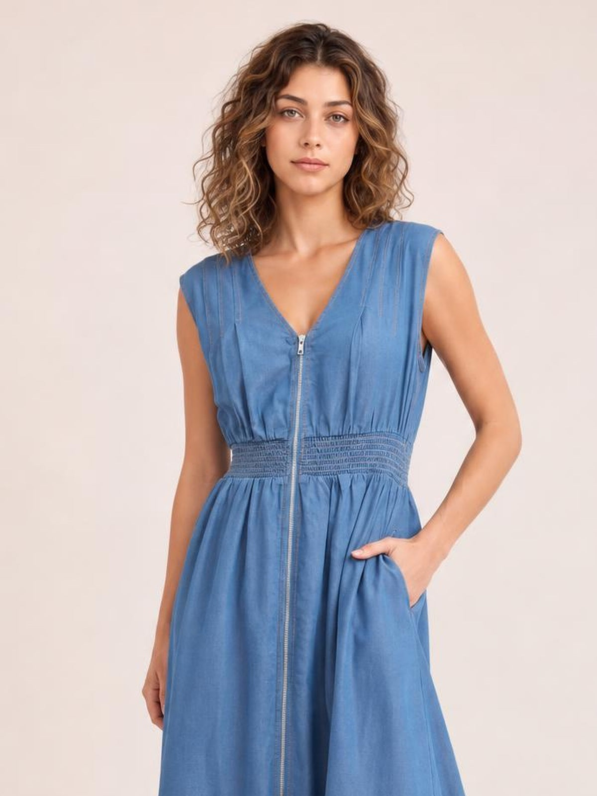 Model poses with one hand in the pocket of a blue denim sleeveless midi dress featuring a V-neck, front zipper, smocked waist, and flared silhouette.