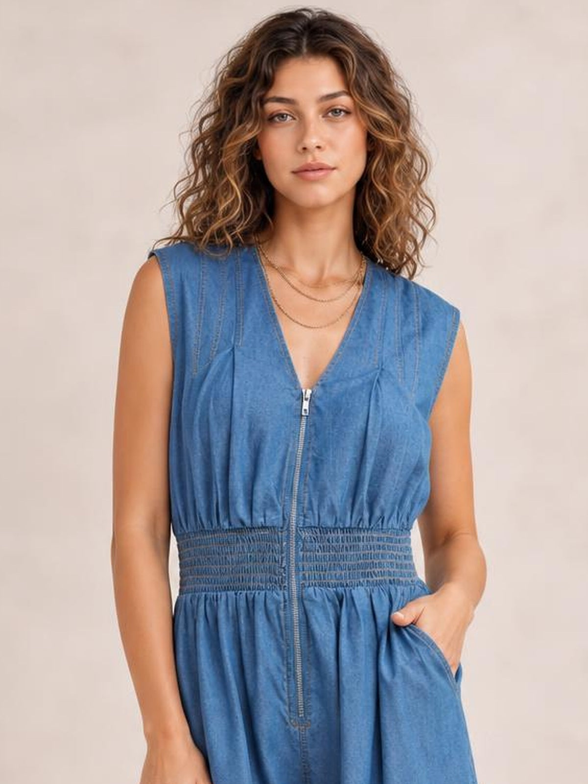 Model poses with one hand in a pocket, wearing a blue denim sleeveless midi dress with a V-neck, front zipper, smocked waist, and gathered skirt.