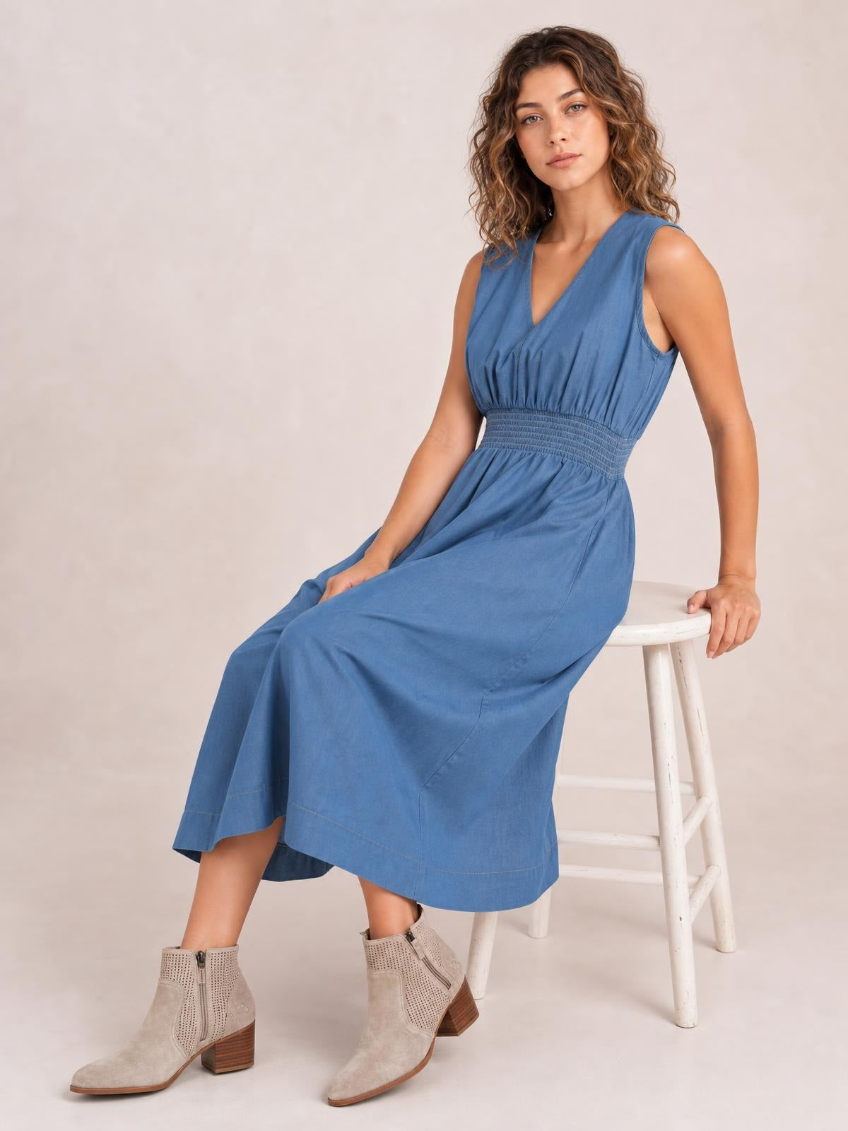 A model sits in a blue denim sleeveless midi dress with a V-neck, gathered bodice, smocked waist, and flowing A-line skirt.