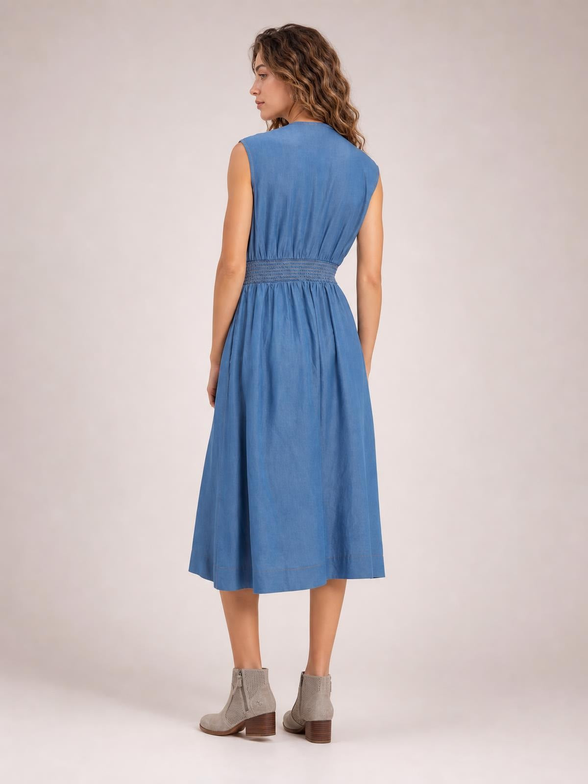Model turned away to show a blue denim sleeveless midi dress with a fitted smocked waist and softly flared, below-knee skirt.