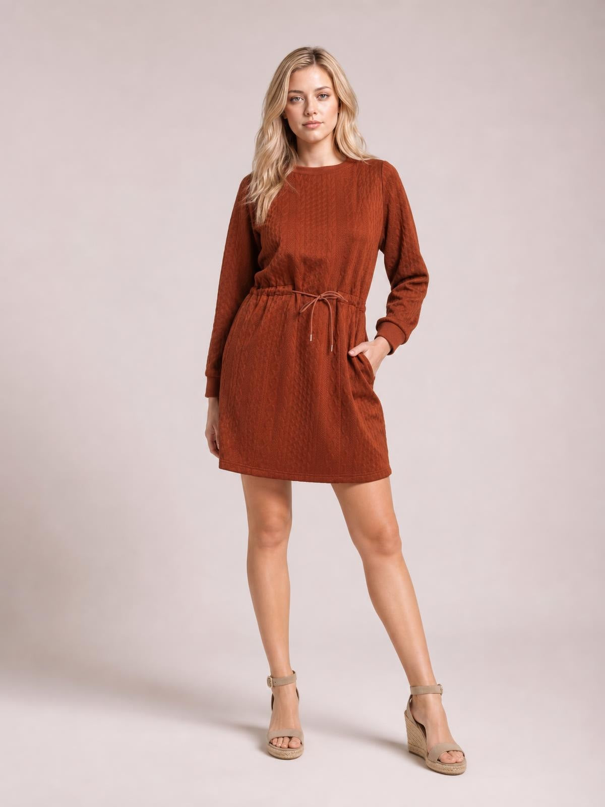 Model poses in a rust brown cable-knit mini dress with long sleeves, a drawstring waist, relaxed silhouette, and side pockets.