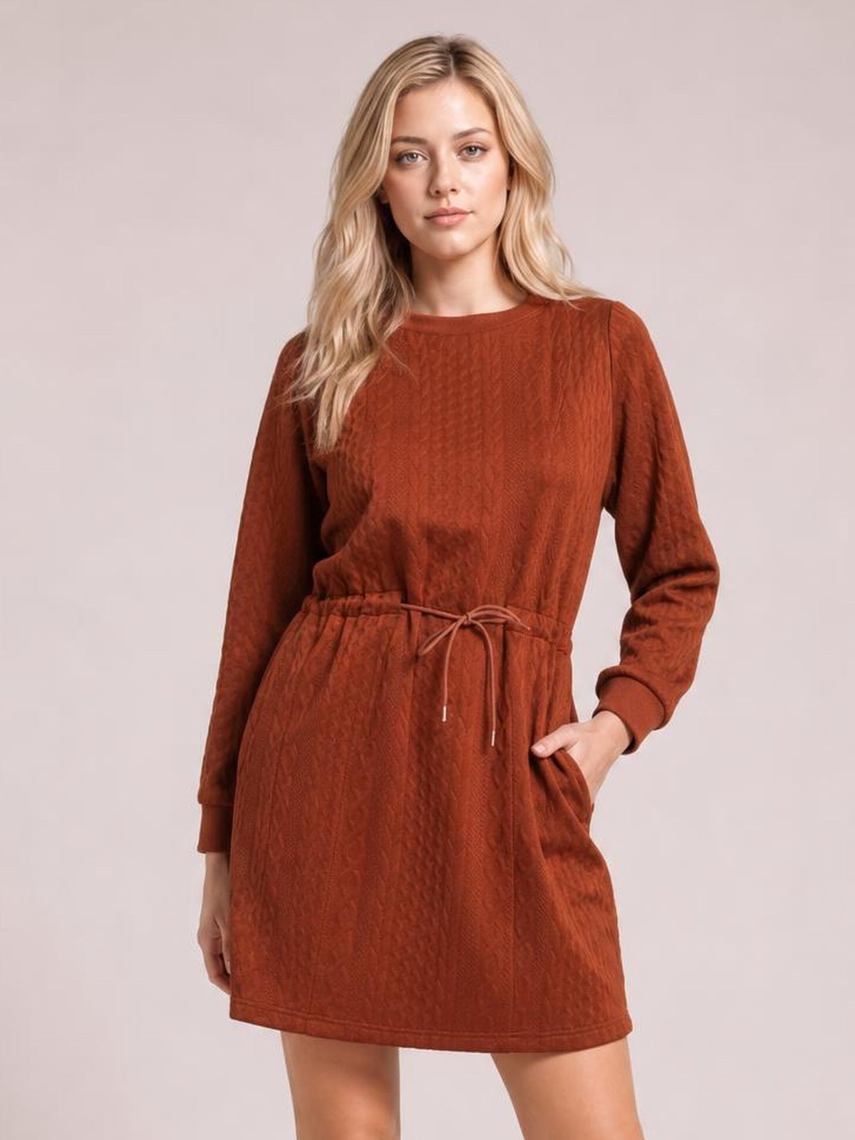 Model wearing a rust-brown cable knit mini dress with long sleeves, a drawstring waist, relaxed A-line silhouette, and side pockets.