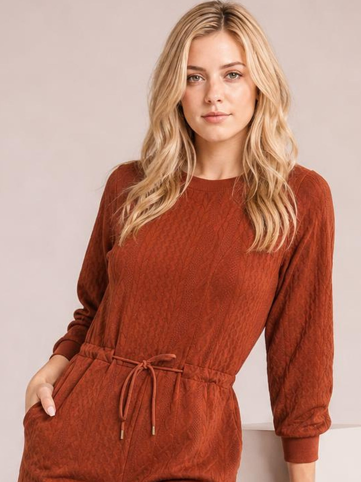 Model wears a rust-brown cable knit mini dress with long sleeves, a crew neckline, drawstring waist, relaxed silhouette, and side pockets.