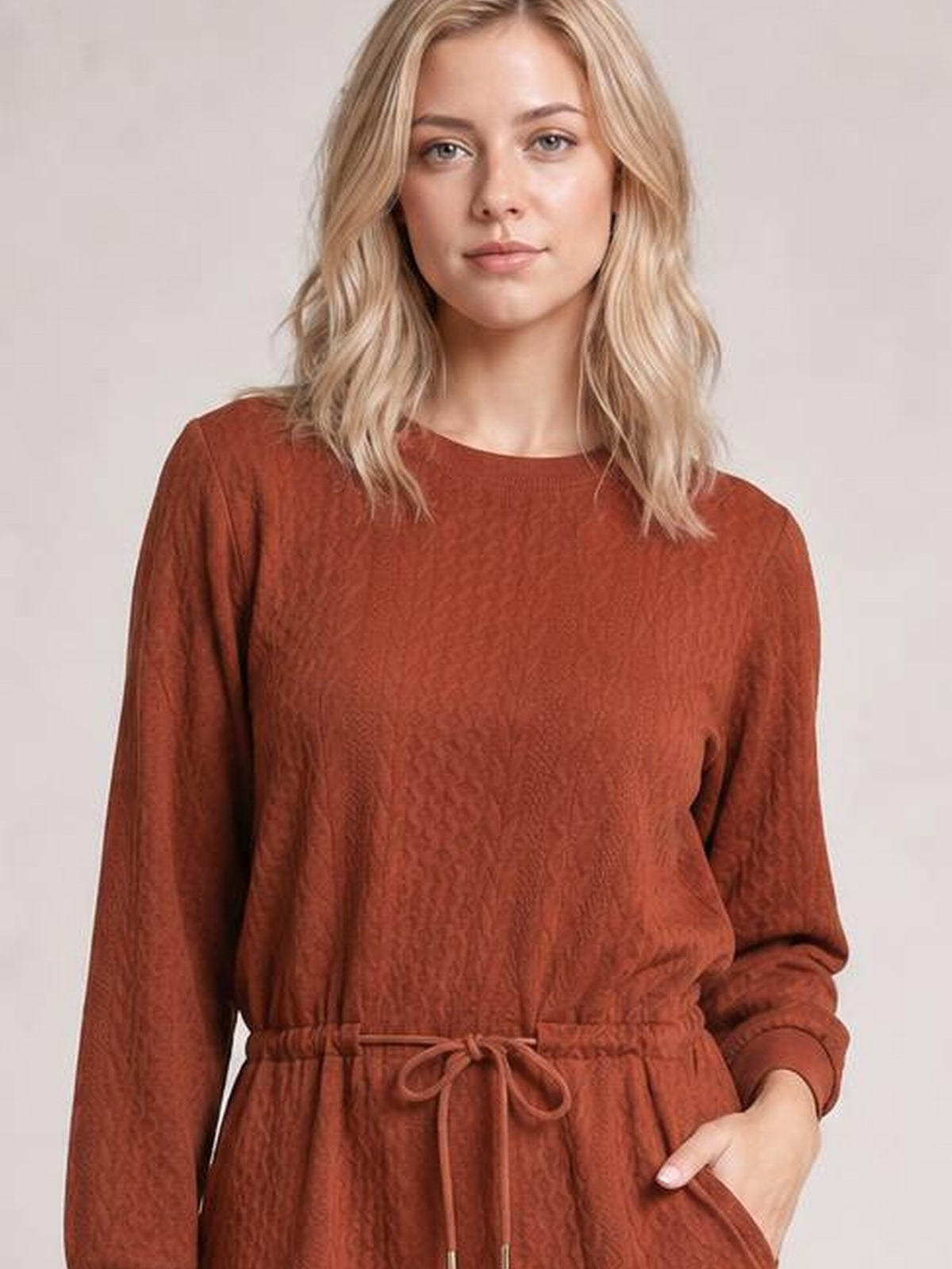 Model wears a rust-brown cable knit mini dress with long sleeves, a drawstring waist, relaxed silhouette, and one visible side pocket.