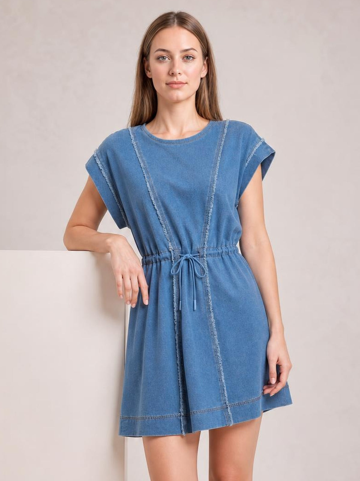 A model stands in a blue denim mini dress with a round neck, cap sleeves, exposed frayed seams, a relaxed silhouette, and adjustable drawstring waist.