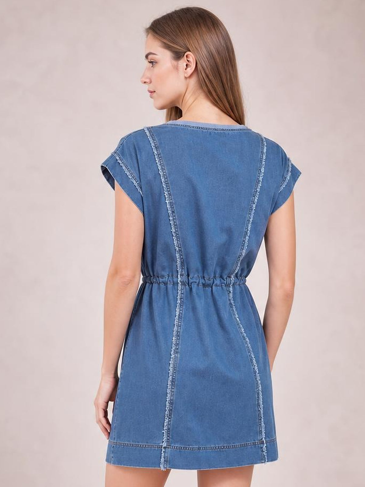 Model shown from behind wearing a blue denim mini dress with cap sleeves, a round neckline, relaxed silhouette, drawstring waist, and frayed exposed seams.