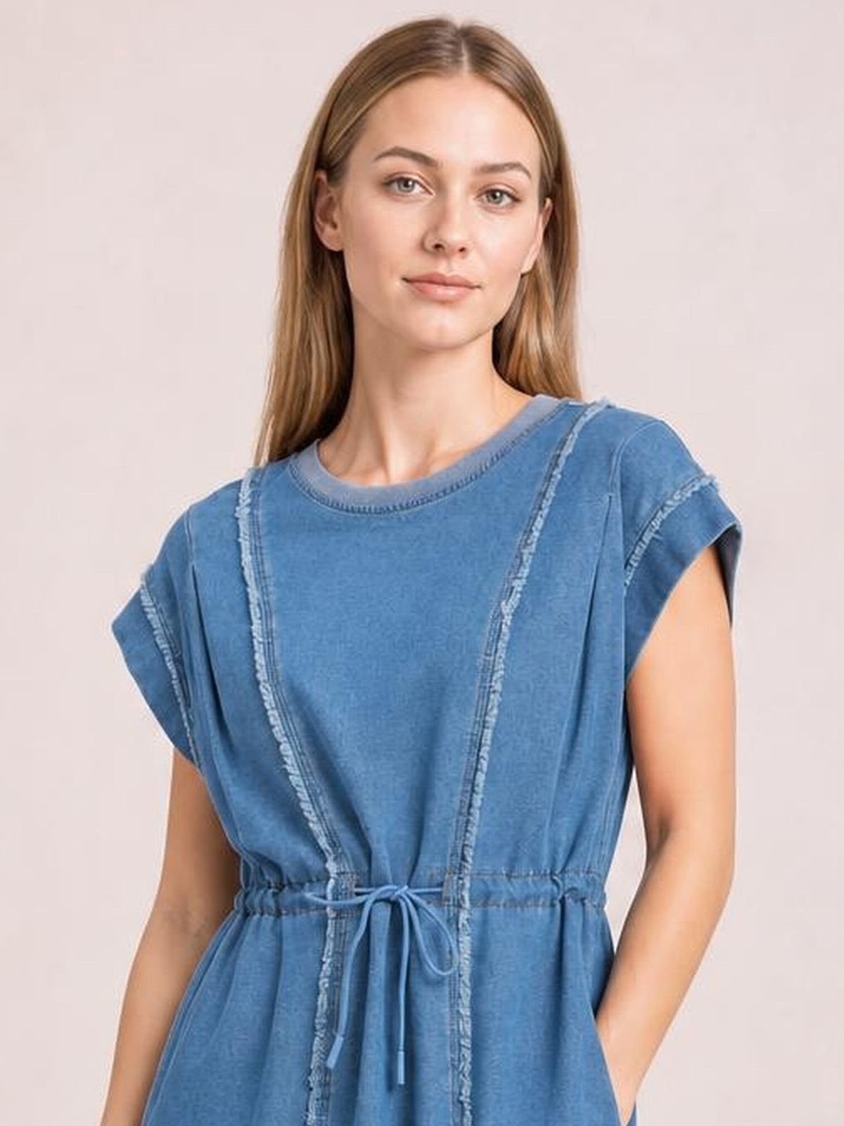Model wearing a blue denim mini dress with a round neck, cap sleeves, frayed exposed seams, relaxed silhouette, drawstring waist, and side pockets.