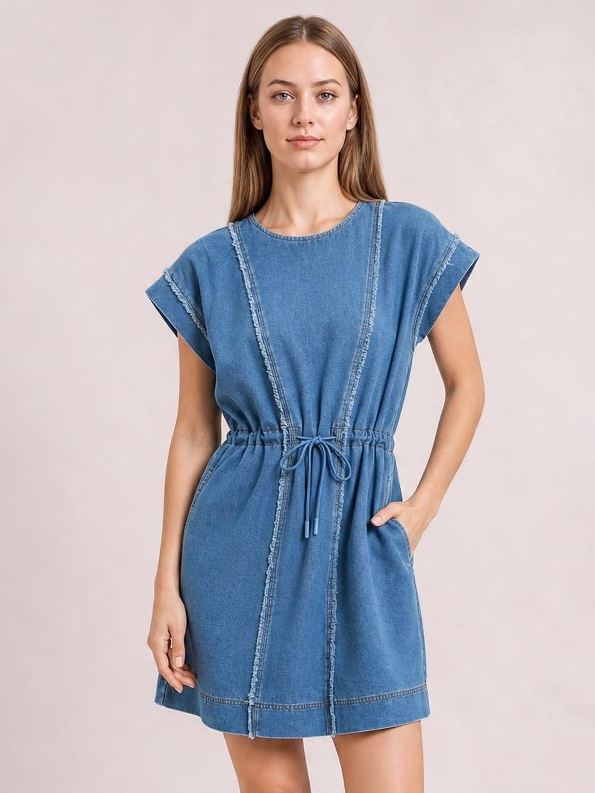 Model poses in a blue denim mini dress with cap sleeves, round neck, relaxed silhouette, drawstring waist, raw frayed seams, and side pockets.