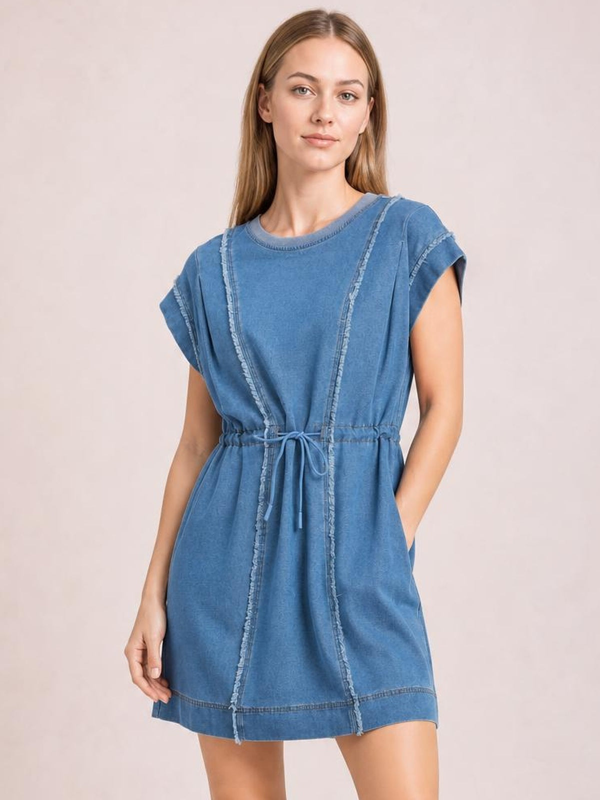 Model wearing a blue denim mini dress with a round neck, cap sleeves, relaxed silhouette, drawstring waist, side pockets, and frayed exposed seams.