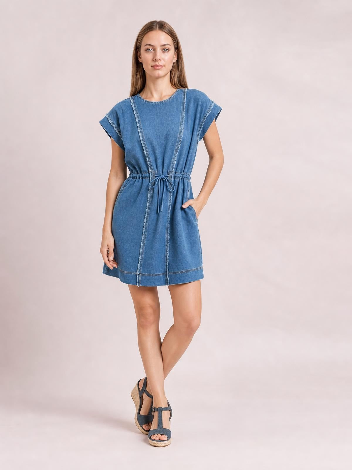 Model poses in a blue denim mini dress with cap sleeves, round neck, frayed exposed seams, adjustable drawstring waist, relaxed silhouette, and side pockets.