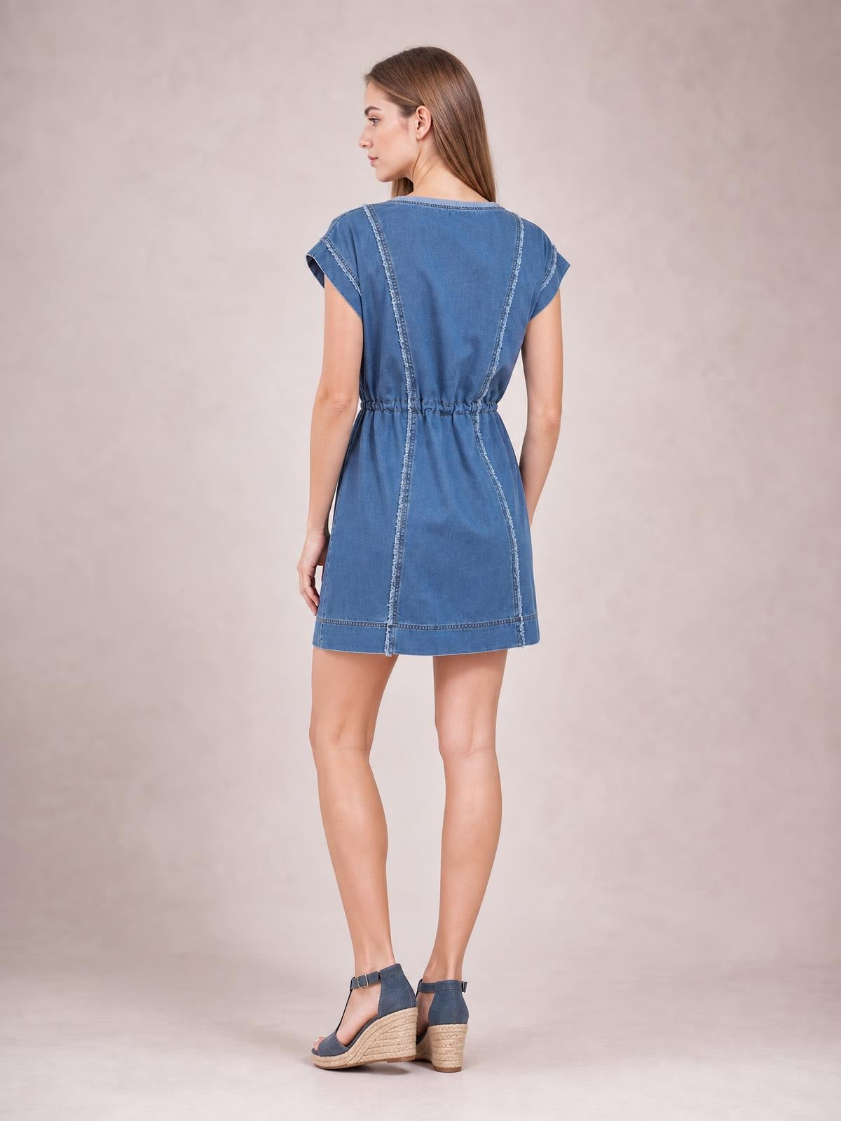 A model stands turned away in a blue denim mini dress with cap sleeves, a drawstring waist, relaxed silhouette, and frayed exposed seams.