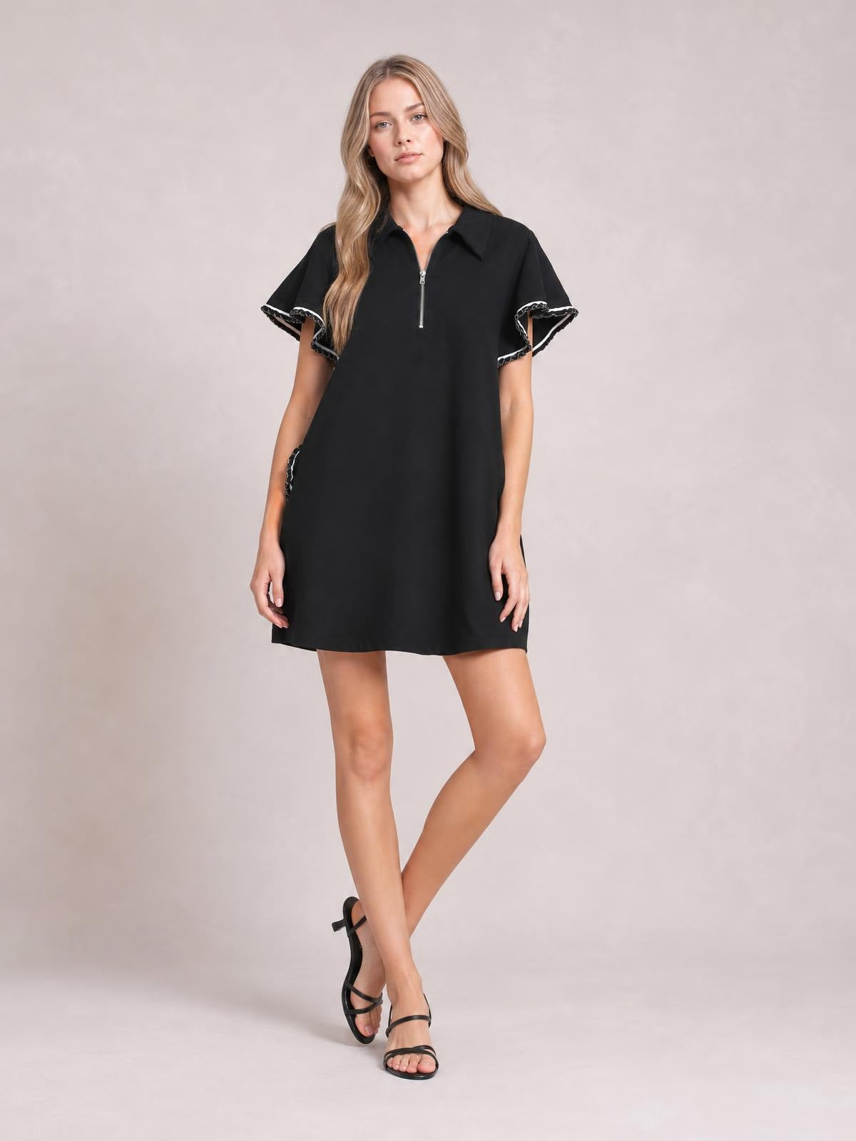 A model poses with one leg crossed in a black mini dress featuring a half-zip collar, ruffled butterfly sleeves with contrast trim, and side pockets.