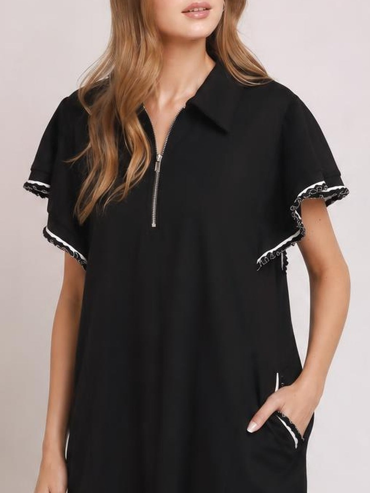 A model wears a black mini dress with a half-zip front, flowing butterfly sleeves trimmed with ruffles, a relaxed silhouette, and a visible side pocket.