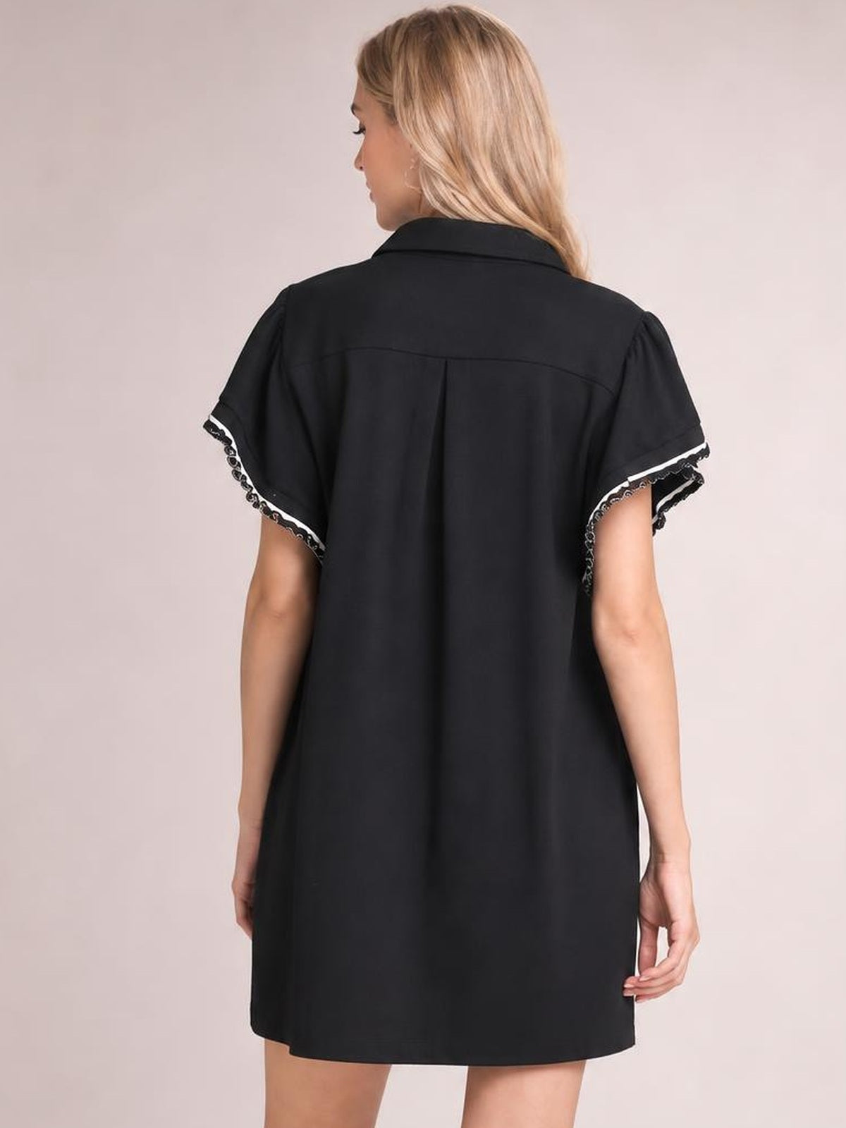 A model stands facing away in a black ruffle mini dress with loose structured silhouette, short butterfly sleeves, contrast trim, and a softly pleated back.