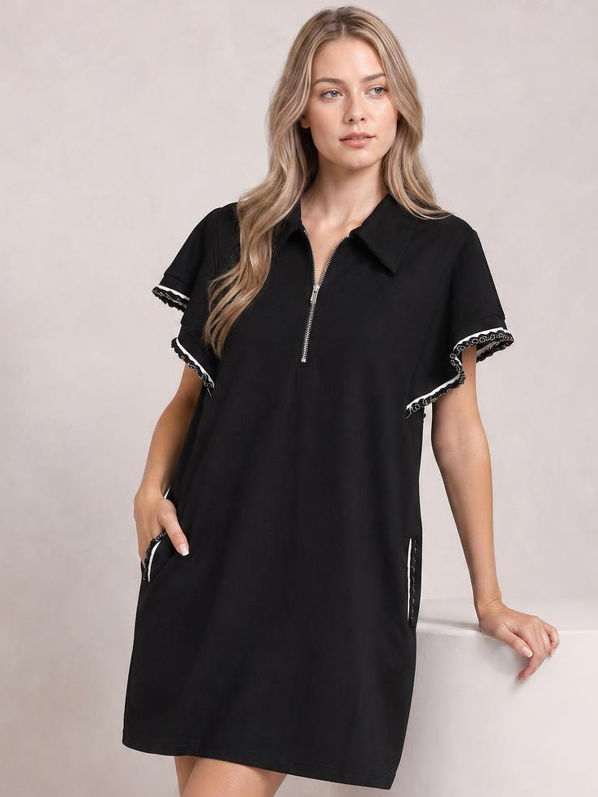 A model wears a black mini dress with a half-zip front, ruffle-trimmed butterfly sleeves, side pockets, and a relaxed straight silhouette, with one hand tucked in a pocket.