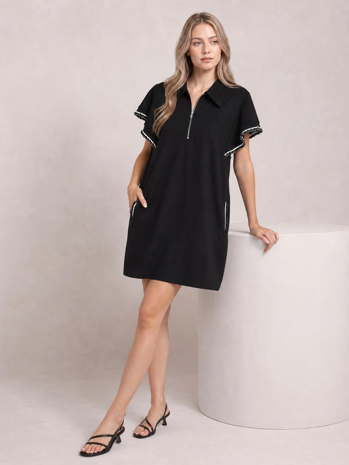 A model poses in a black mini dress with a half-zip collared front, ruffled short sleeves, contrast trim, straight silhouette, and side pockets.