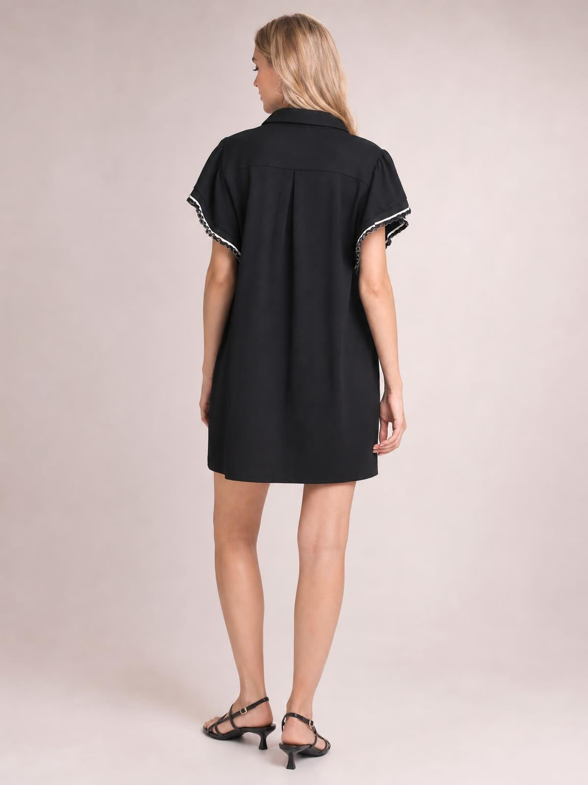 Model turned away wears a black mini dress with a relaxed straight silhouette, collared neckline, short butterfly sleeves, and ruffled contrast trim.