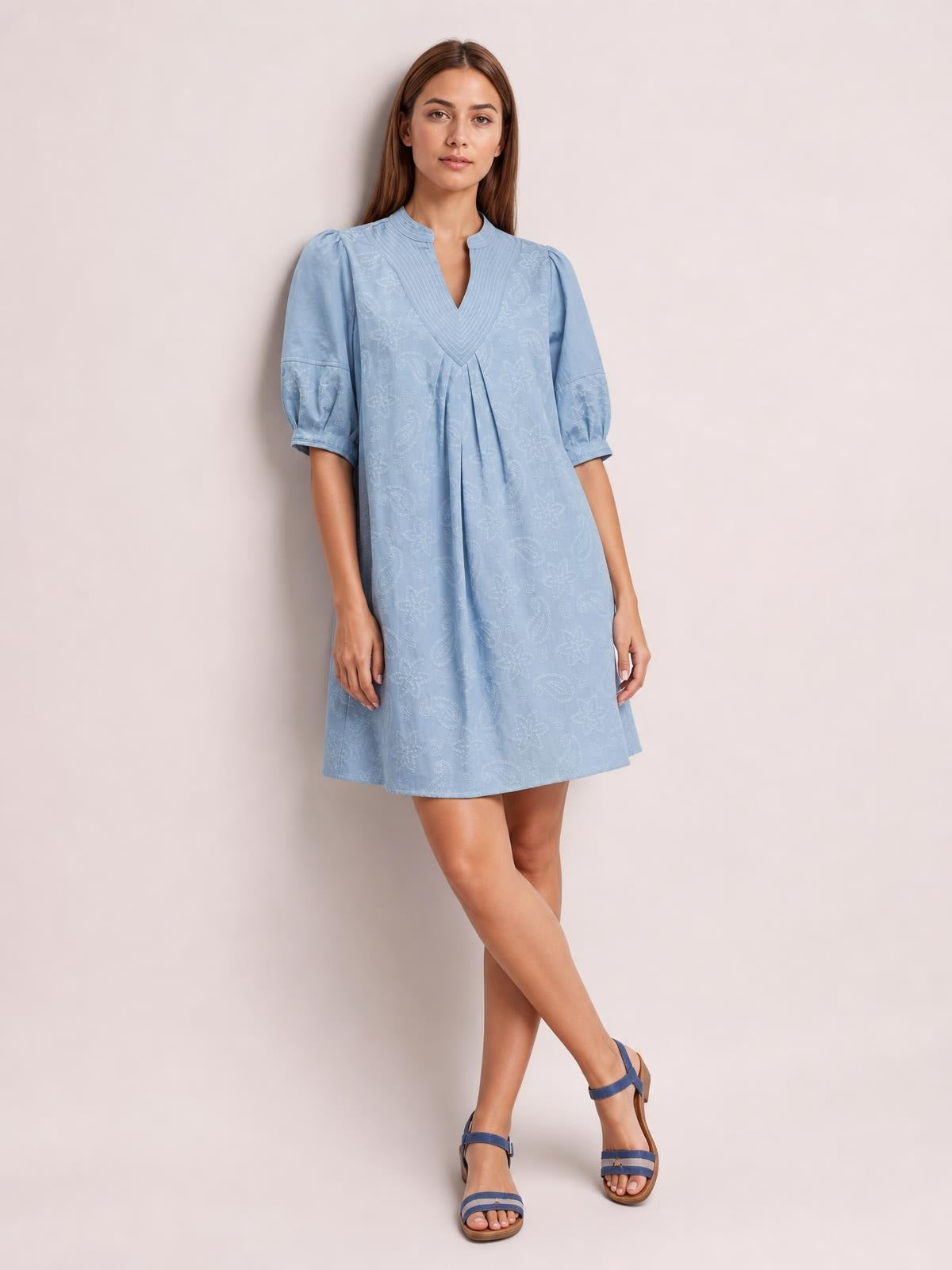 A model stands with crossed legs in a light blue printed denim dress featuring contrast stitching, a split V-neck, pintuck detail, puff sleeves, pleats, and a relaxed silhouette.