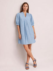A model stands with crossed legs in a light blue printed denim dress featuring contrast stitching, a split V-neck, pintuck detail, puff sleeves, pleats, and a relaxed silhouette.