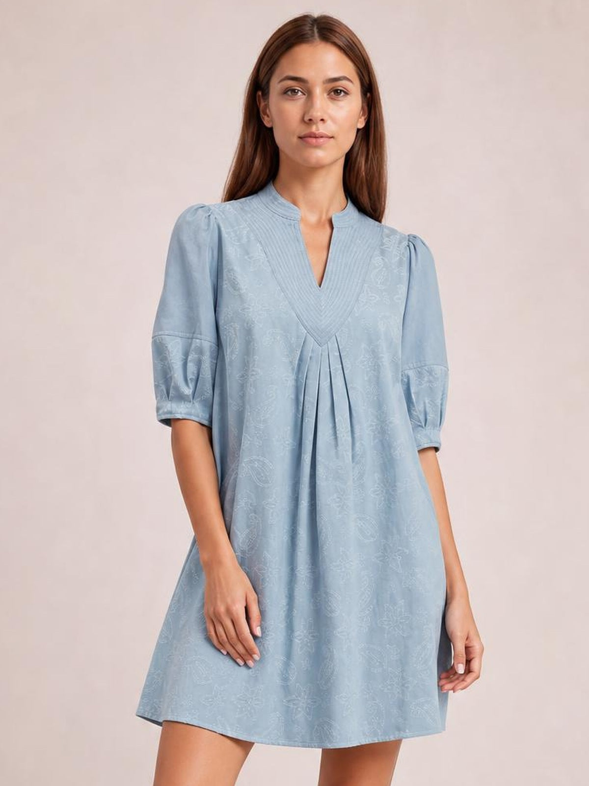 A model stands facing forward in a light blue printed denim dress with a split V-neck, pintuck collar, contrast stitching, puff sleeves, front pleats, and a relaxed silhouette.