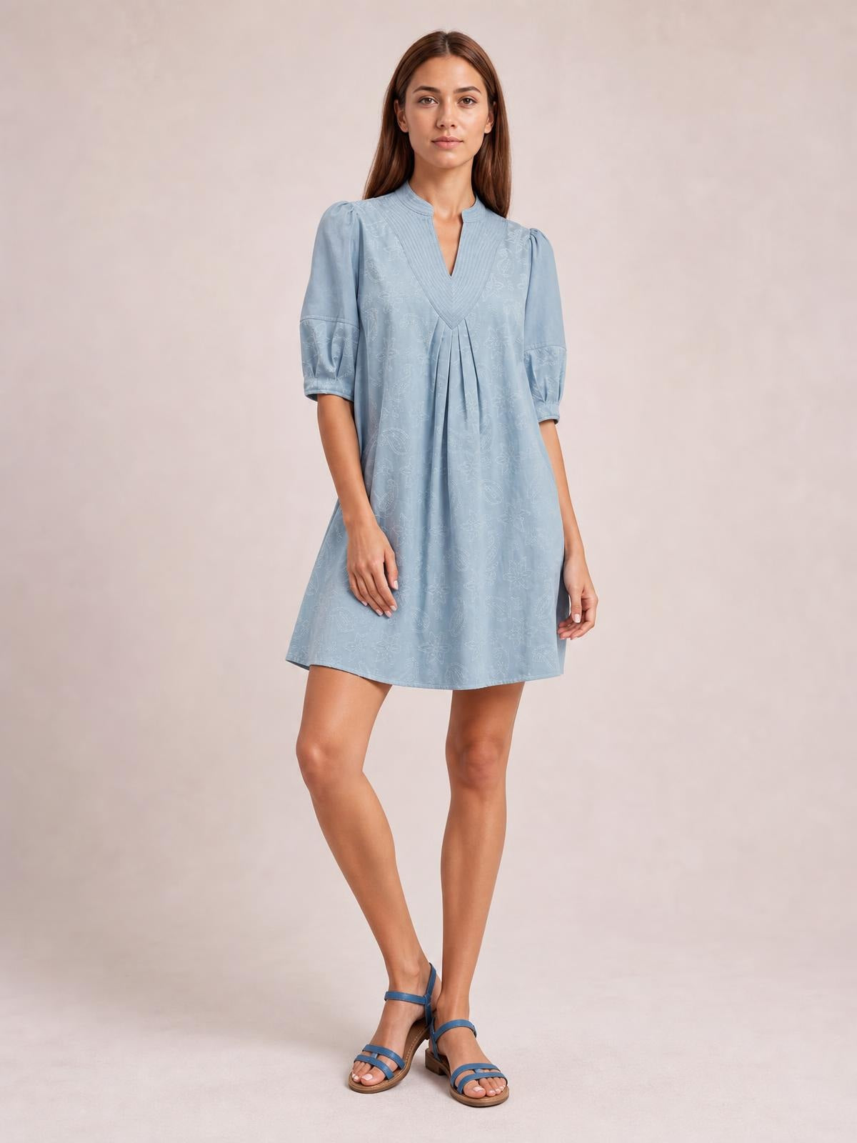 A model poses in a light blue printed denim dress with a split V-neck, pintuck collar, pleated front, elbow-length puff sleeves, and a relaxed A-line silhouette.