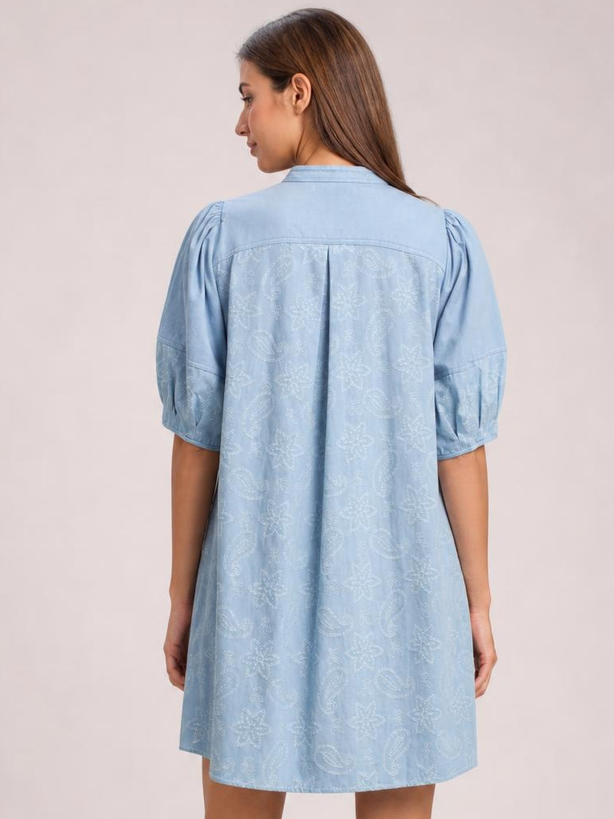 Model shown from behind wearing a light blue printed denim dress with contrast stitching, elbow-length puff sleeves, a back yoke pleat, and a relaxed silhouette.