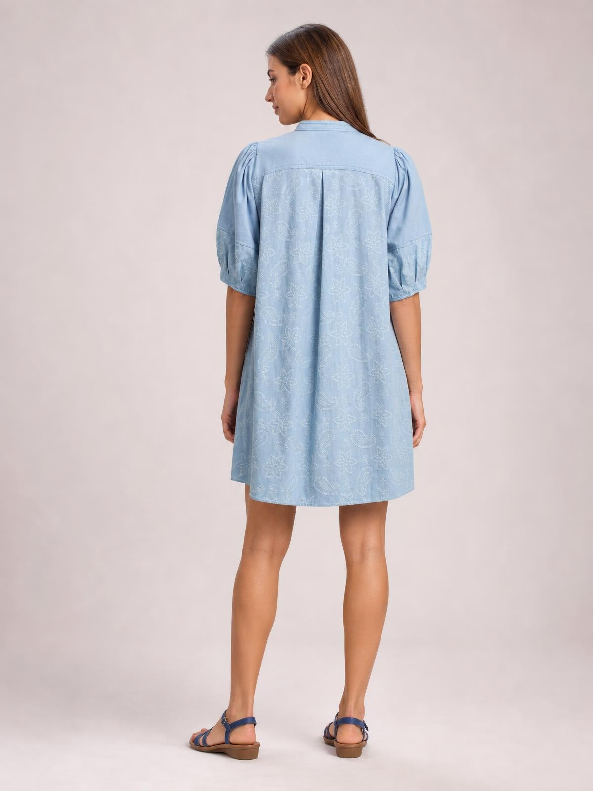Model viewed from the back in a light blue printed denim dress with contrast stitching, elbow-length puff sleeves, pleated detailing, and a relaxed knee-length silhouette.