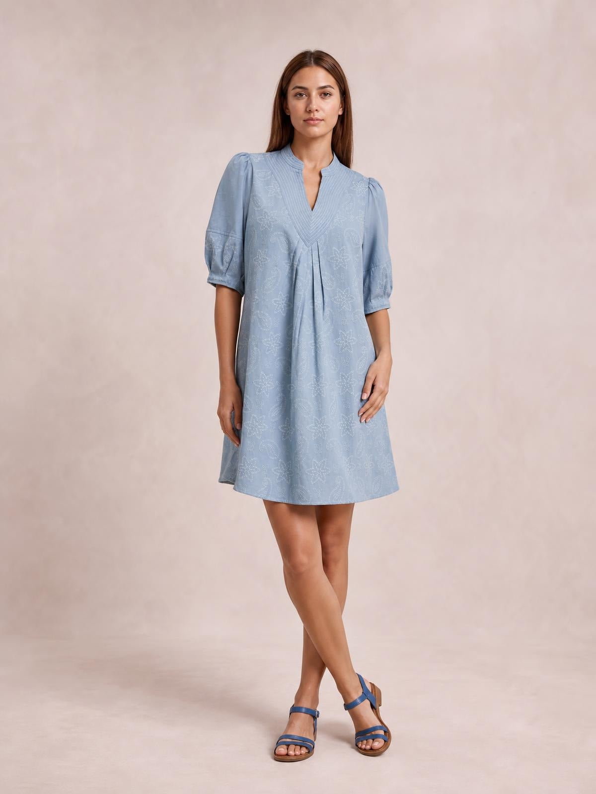 Model poses in a light blue printed denim dress with a split V-neck, pintuck collar, elbow-length puff sleeves, pleated front, contrast stitching, and a relaxed silhouette.