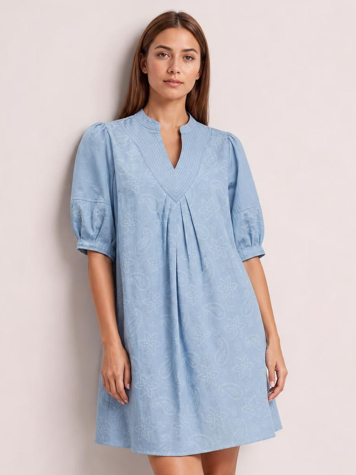 A model stands in a light blue printed denim dress with a split V-neck, pintuck collar, contrast stitching, puff sleeves, pleated front, and relaxed silhouette.
