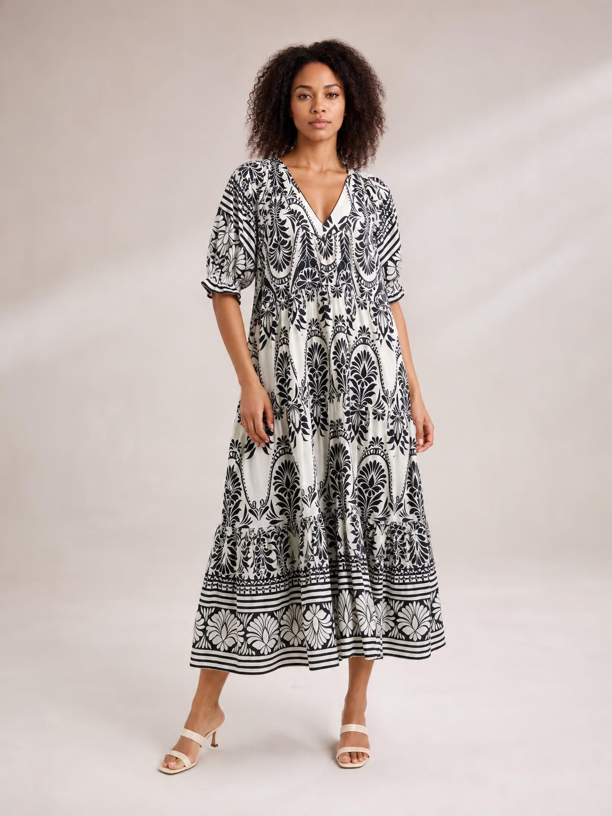 Model stands in a flowing black-and-white two-tone border print midi dress with a V-neck, puff sleeves, tiered silhouette, and patterned hem.