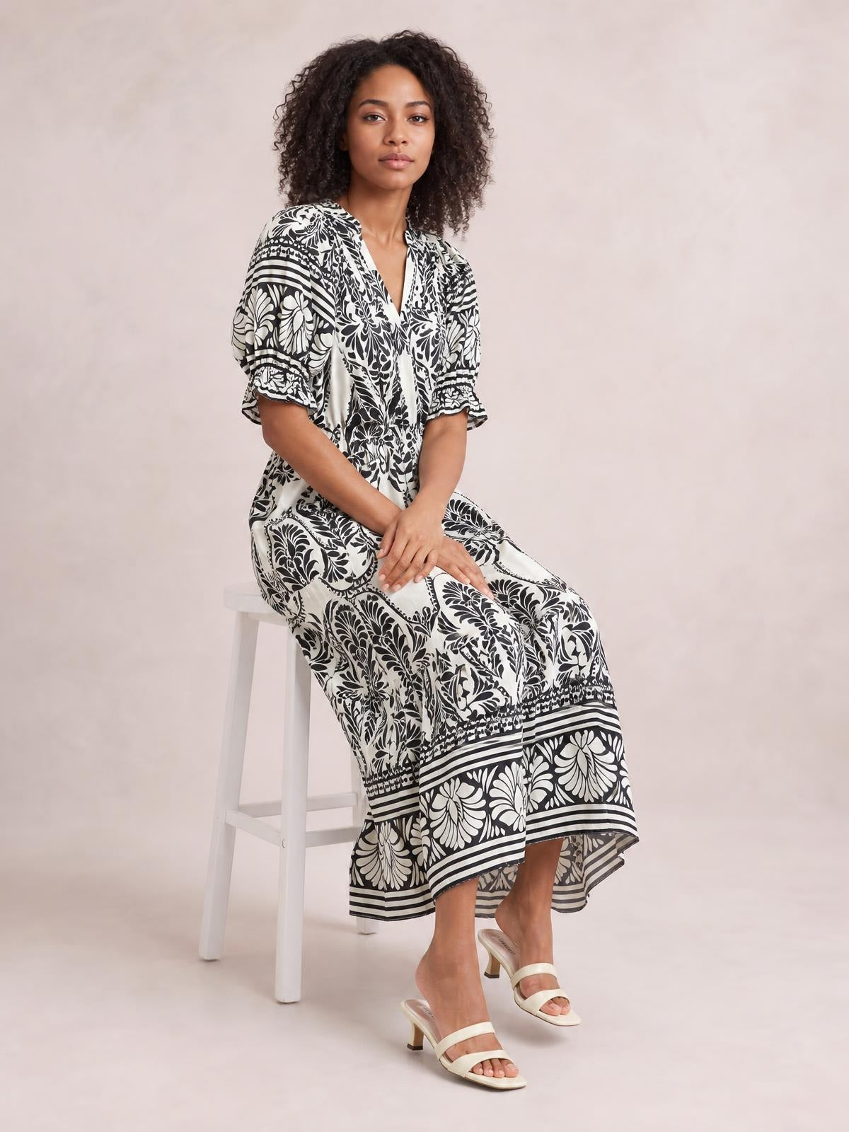 Model seated in a black-and-white two-tone border print midi dress with a V-neck, flutter sleeves, flowy tiered silhouette, and floral detailing.